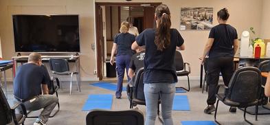 Des personnes en salle réalisant un exercice debout devant des chaises.