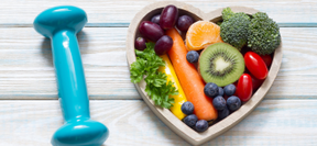 Haltère bleu posé à côté d’un bol en forme de cœur rempli de fruits et légumes frais sur un fond en bois clair.