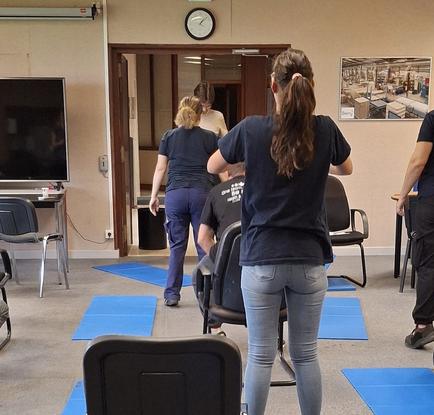 Des personnes en salle réalisant un exercice debout devant des chaises.